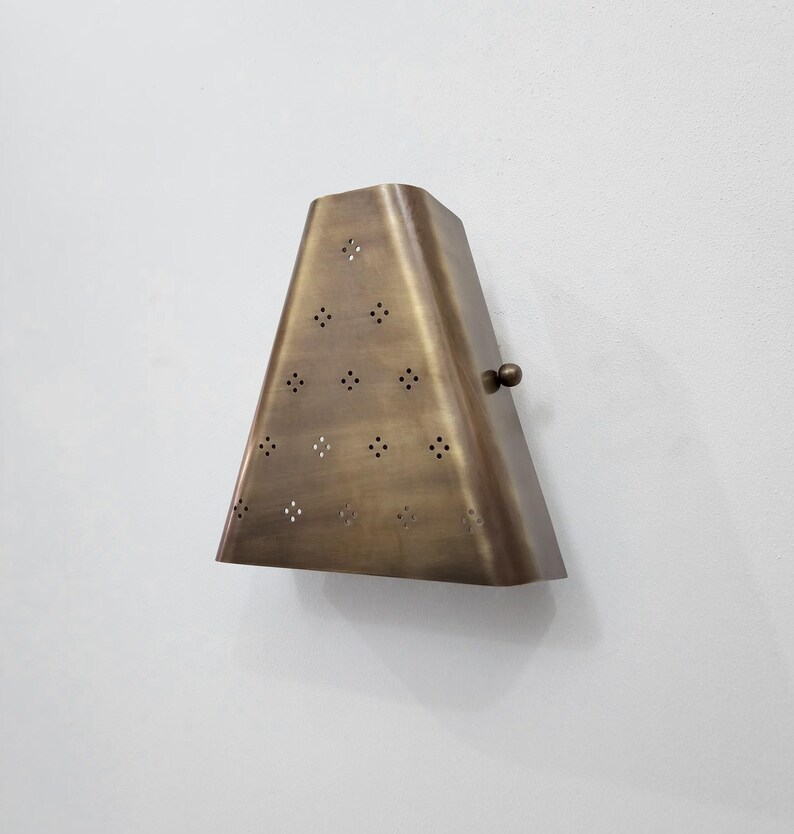 May include: A brass-colored wall sconce with a trapezoidal shape. The sconce has a series of small, evenly spaced holes and a small, round knob on the side. It is mounted on a white wall.
