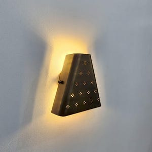 May include: A bronze-colored wall sconce with a triangular shape. The sconce has a series of small, decorative holes arranged in a pattern. The light from the sconce illuminates the wall behind it, creating a warm glow. The sconce is mounted on a white wall.