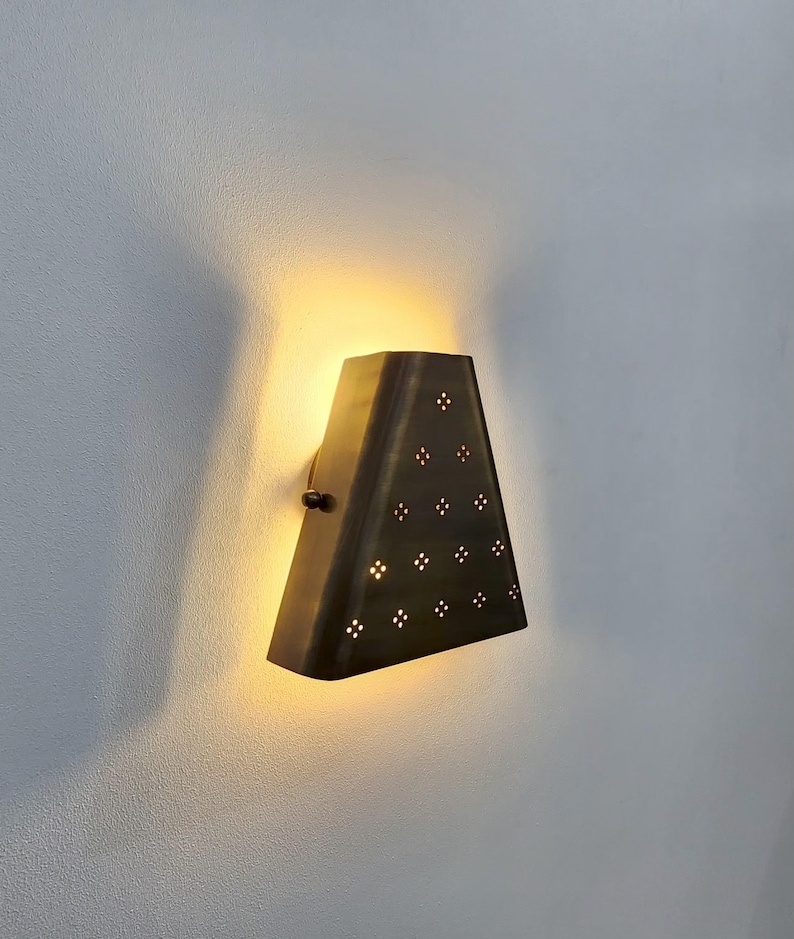 May include: A bronze-colored wall sconce with a triangular shape. The sconce has a series of small, decorative holes arranged in a pattern. The light from the sconce illuminates the wall behind it, creating a warm glow. The sconce is mounted on a white wall.