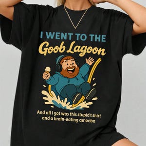 May include: Black t-shirt with the text "I WENT TO THE Goob Lagoon" in teal and yellow. The shirt features a cartoon illustration of a man with headphones, an ice cream cone, and the text "And all I got was this stupid t-shirt and a brain-eating amoeba."