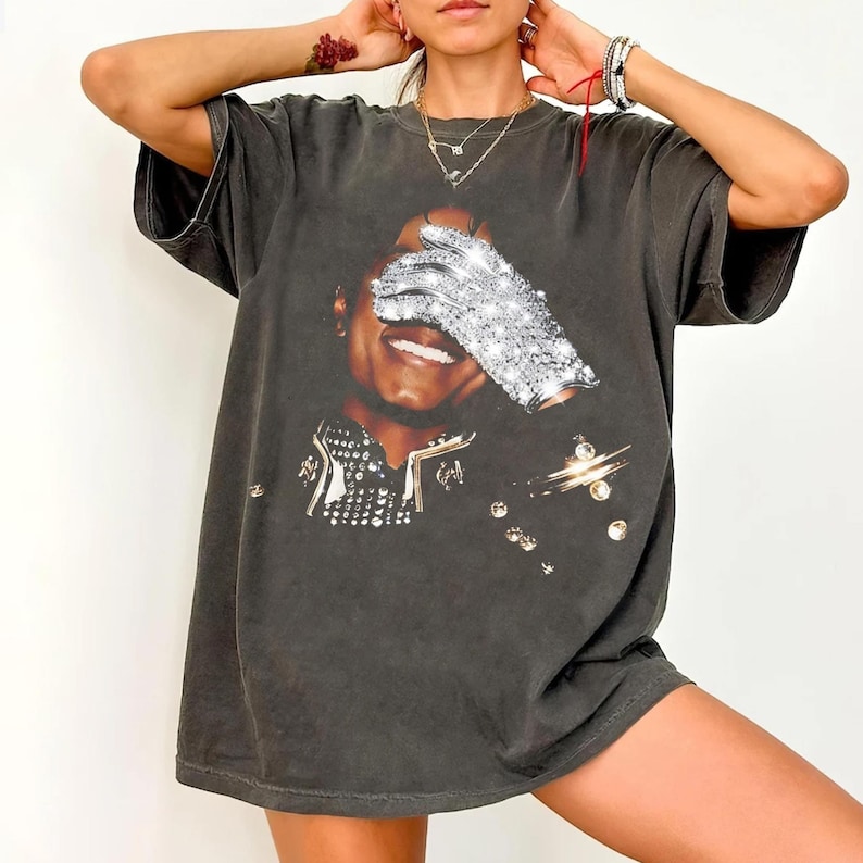 K&ouml;nnte beinhalten: Dunkelgraues T-Shirt mit einem Motiv eines l&auml;chelnden Gesichts und eines funkelnden Handschuhs, der die Augen bedeckt. Das T-Shirt hat einen Vintage-Look und glitzernde Akzente. Ein l&auml;ssiges Kleidungsst&uuml;ck.