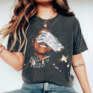 K&ouml;nnte beinhalten: Dunkelgraues T-Shirt mit einer Grafik einer l&auml;chelnden Person mit einem funkelnden Handschuh &uuml;ber den Augen. Das Shirt hat einen Vintage-Look und ist mit funkelnden Verzierungen versehen. Eine goldene Halskette und ein Armband sind ebenfalls sichtbar.