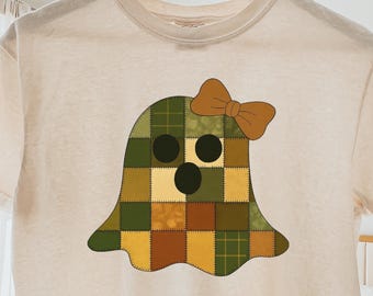 Pode incluir: T-shirt creme com um design de fantasma de patchwork e um laço castanho. O fantasma é composto por quadrados verdes, castanhos e amarelos. A camisa está pendurada em um cabide de madeira.