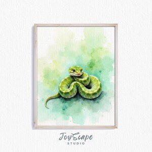 Puede incluir: Pintura a la acuarela de una serpiente verde enroscada con una cara sonriente. La serpiente se encuentra sobre un fondo suave de acuarela verde. La obra de arte está enmarcada en un marco de madera clara. El texto "JoyScape Studio" está en la parte inferior.
