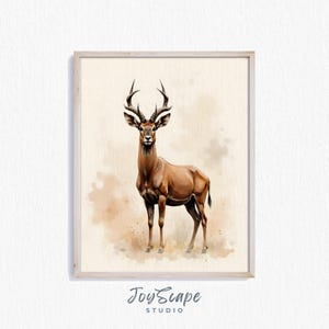 May include: A framed watercolour painting of a brown antelope with prominent, curved horns. The artwork is set against a neutral background with earthy tones. The painting is enclosed in a light-coloured wooden frame. The artist's signature, "JoyScape Studio," is visible.