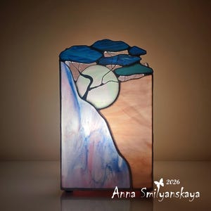 Puede incluir: Obra de arte en vidriera que representa un paisaje con una gran luna, árboles y nubes. La obra presenta tonos azules, rosas y blancos, con el nombre del artista y el año visibles. La pieza es rectangular y se mantiene en posición vertical.