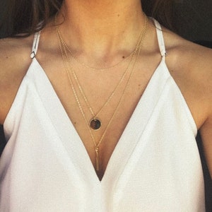 Puede incluir: Un collar de oro de varios niveles con un pequeño colgante redondo y un colgante largo y delgado.