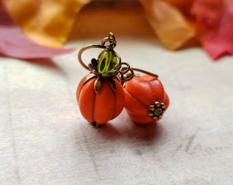 May include: A pair of orange pumpkin-shaped earrings with green leaves and gold accents. The earrings are on a white surface.