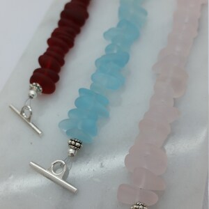 Pu&ograve; includere: Tre braccialetti in vetro di mare con chiusure a scorrimento argentate. I braccialetti sono realizzati in vetro di mare rosso, blu e rosa.