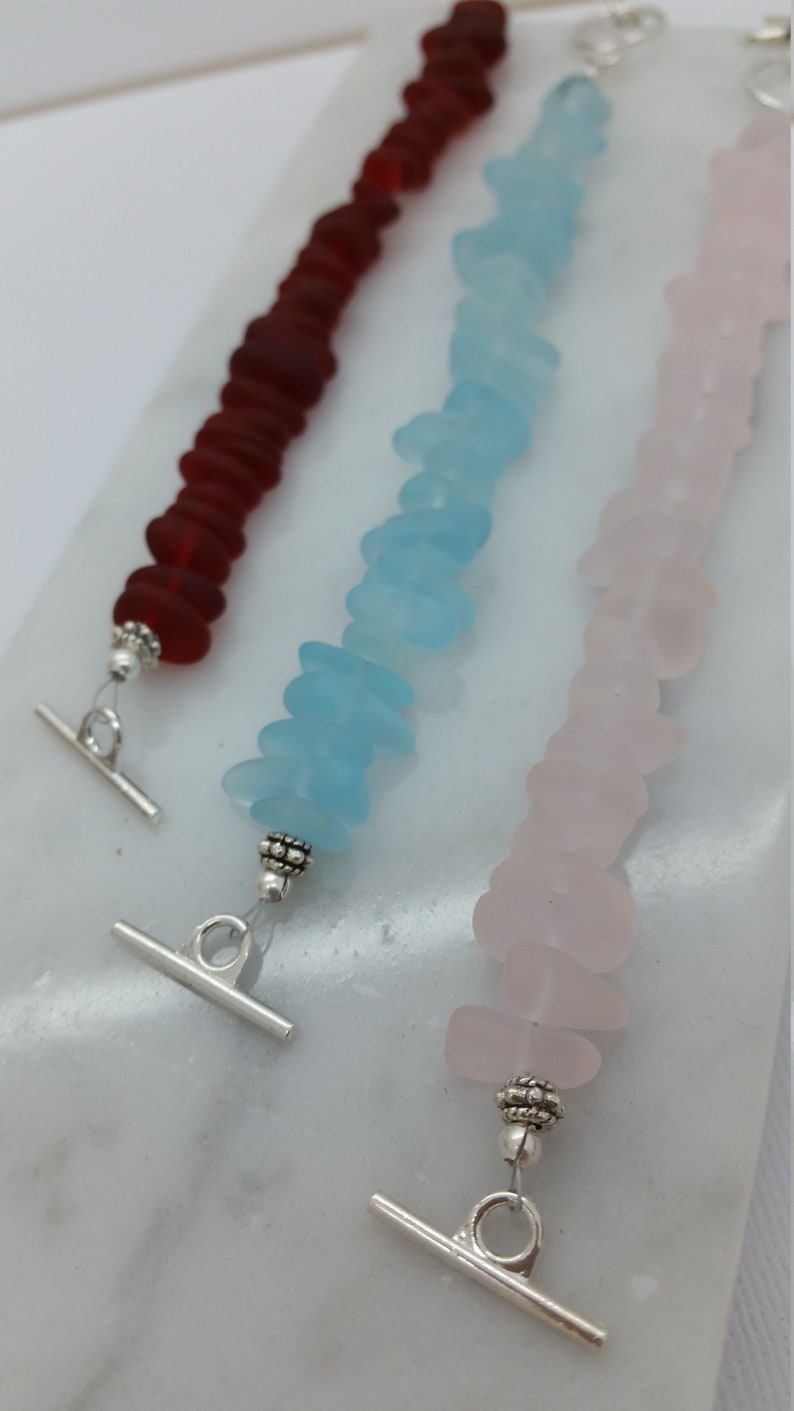 Pu&ograve; includere: Tre braccialetti in vetro di mare con chiusure a scorrimento argentate. I braccialetti sono realizzati in vetro di mare rosso, blu e rosa.