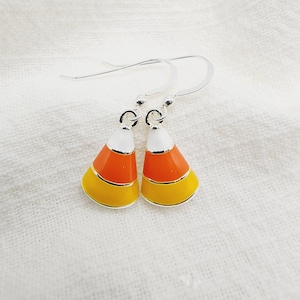 May include: A pair of candy corn earrings. Each earring features a silver hook and a candy corn design with white, orange, and yellow enamel. The earrings are set against a white fabric background.