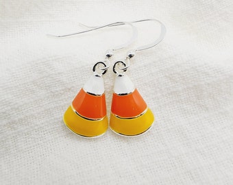 Puede incluir: Un par de pendientes de caramelo de maíz. Cada pendiente presenta un gancho plateado y un diseño de caramelo de maíz con esmalte blanco, naranja y amarillo. Los pendientes están colocados sobre un fondo de tela blanca.