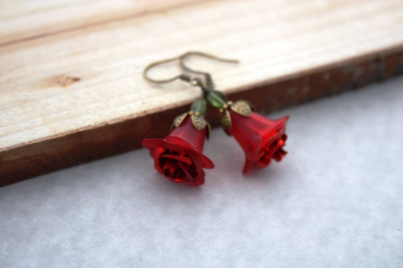 May include: A pair of red rose earrings with green accents. The earrings are made of glass and have a bell shape.