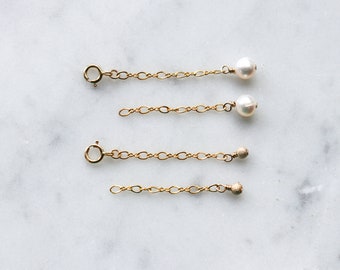 May include: Four gold-colored chain extenders. Two have a small white pearl at the end, and two have a small textured gold bead. Each extender has a clasp at one end and a decorative element at the other.