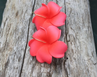 Puede incluir: Un par de pendientes de flores rosas con aros dorados. Los pendientes están hechos de tela y tienen forma de flores de frangipani.