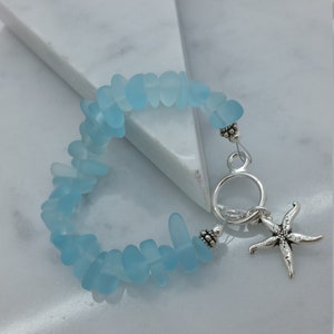 Pu&ograve; includere: Un braccialetto in vetro di mare blu chiaro con un ciondolo a forma di stella marina argentata e una chiusura argentata.