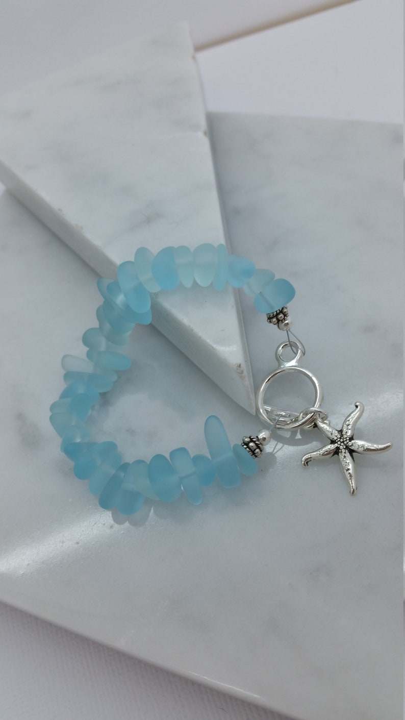 Pu&ograve; includere: Un braccialetto in vetro di mare blu chiaro con un ciondolo a forma di stella marina argentata e una chiusura argentata.
