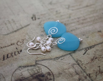 May include: A pair of earrings featuring light blue, heart-shaped stones with silver wire swirls. Small white pearls dangle below the blue stones. The earrings are set against a vintage-style background.
