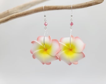 Puede incluir: Un par de pendientes florales con pétalos blancos y amarillos con bordes rosas. Cada pendiente tiene un gancho plateado, una cuenta de cristal rosa y un alambre que suspende la flor. Fondo claro.