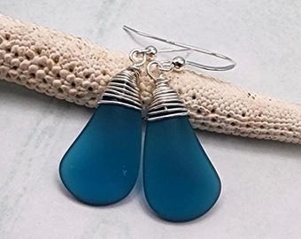 May include: A pair of teal teardrop earrings. The earrings feature a smooth, rounded teal glass pendant wrapped with silver wire. The ear wires are silver. The earrings are displayed on a textured, off-white surface.