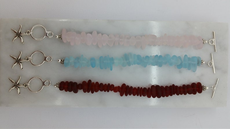 Pu&ograve; includere: Tre braccialetti in vetro di mare con ciondoli a forma di stella marina argentati e chiusure a scorrimento. I braccialetti sono realizzati in vetro di mare rosa, blu e rosso.