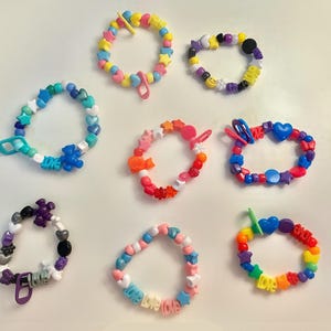 May include: A collection of eight colorful beaded bracelets featuring various shapes including stars, hearts, bears, and letter beads spelling love. The bracelets are constructed with elastic cord and include assorted plastic charms in shades of pink, blue, yellow, orange, purple, and black.
