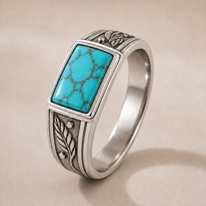 May include: A silver-toned ring featuring a rectangular turquoise stone with dark brown veining set in the center. The band is crafted with an oxidized finish and displays detailed engravings of leaves and small circular accents along the sides.