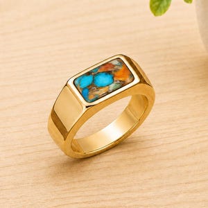 May include: A gold-toned ring featuring a rectangular face inlaid with a stone mosaic. The inlay displays a vibrant pattern of bright blue, orange, and brown flecks. The band is smooth, polished, and has a thick, solid construction.
