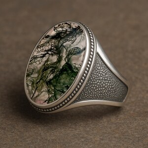 May include: A silver ring with an oval-shaped moss agate stone. The stone features green and black patterns resembling a landscape. The ring has a textured band and a beaded border around the stone.
