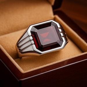 May include: A silver ring featuring a large, rectangular, deep red gemstone. The ring has a thick band with three parallel grooves on each side of the gemstone. The ring is presented in a brown jewellery box with a tan interior.