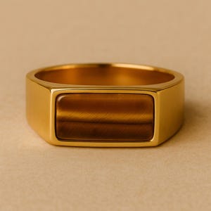 May include: A gold-colored signet ring with a rectangular tiger's eye stone. The ring has a polished finish and a classic, masculine design. The stone is a warm brown color with natural striations.