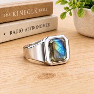 May include: A silver-toned ring featuring a rectangular, faceted labradorite gemstone with iridescent blue and gold flashes. The stone is set in a polished metal bezel, showcasing a smooth, wide band that tapers toward the back of the ring.