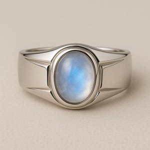 May include: A silver ring featuring an oval, light blue gemstone. The ring has a polished finish and a contemporary design. The gemstone is set in a bezel setting, and the band has a geometric design on the sides.