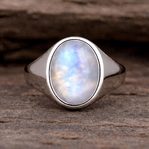 May include: A silver ring featuring an oval moonstone with an iridescent sheen. The moonstone displays a milky white base with blue and yellow hues. The ring has a polished band, and the stone is set in a bezel setting.