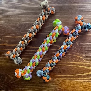 May include: Three bone-shaped dog toys made from braided rope. The toys feature a mix of colours, including brown, orange, green, purple, and blue. They are arranged on a dark wooden surface.
