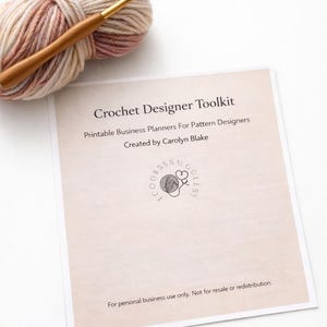 May include: A ball of yarn in shades of beige, pink, and brown with a wooden crochet hook. A printed sheet of paper reads "Crochet Designer Toolkit" and "Printable Business Planners For Pattern Designers".