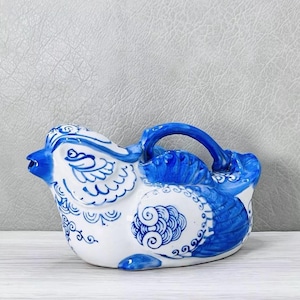 May include: A ceramic bird-shaped pitcher in white and blue. The pitcher features a detailed design with blue floral patterns and a handle. The spout is blue, and the overall design is reminiscent of traditional pottery.