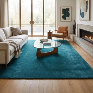 May include: A large, solid teal area rug with a plush, soft texture covering a significant portion of the light-coloured wooden floor in a living room. The rug features a deep, uniform blue-green colour and a rectangular shape with clean edges.