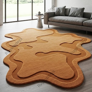 May include: A large, abstract rug in shades of brown and tan. The rug has a layered, organic design with a soft, textured surface. The rug is placed on a wooden floor in a living room setting.