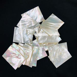 May include: A collection of iridescent, square-shaped mother-of-pearl tiles. The tiles have a pearlescent sheen with hints of pink, green, and blue. They are scattered against a black background, showcasing their natural beauty and texture.