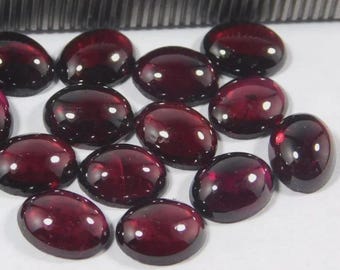 May include: A collection of oval-shaped, deep red gemstones. The polished stones have a glossy surface, reflecting light. The gems are arranged on a white surface, with a metal object in the background. These could be used for jewelry making.