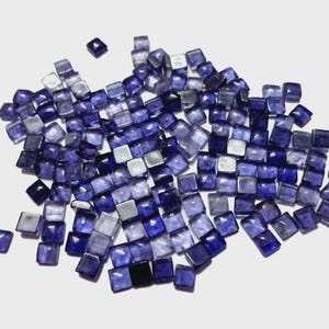 Puede incluir: Un conjunto de pequeñas baldosas cuadradas de vidrio azul y morado dispersas sobre una superficie blanca. Las baldosas varían en tonalidad, algunas parecen más translúcidas. Estas baldosas decorativas son ideales para mosaicos y manualidades.