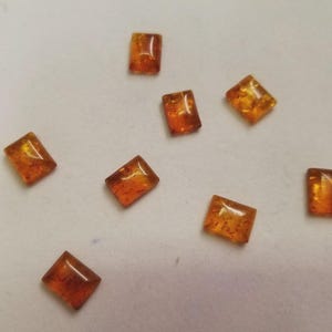 May include: Eight amber-coloured, rectangular gemstones are scattered on a white surface. The polished stones have a translucent appearance, with variations in colour and internal patterns. They are suitable for jewellery making.
