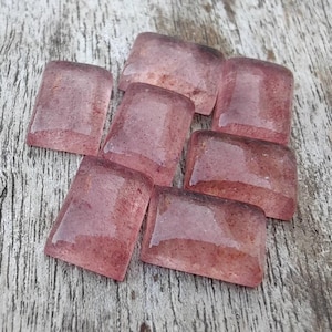 May include: Seven rectangular, translucent, pink gemstones with a slightly rounded shape. The stones have a speckled appearance and are arranged on a light-coloured, textured wooden surface. The gemstones are likely for jewellery making or crafting.