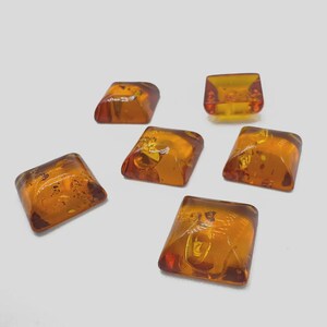 May include: Six amber-colored, square-shaped gemstones with rounded edges are scattered on a white surface. The gems have a translucent quality, with variations in color from light yellow to deep orange. The gems appear to be polished.