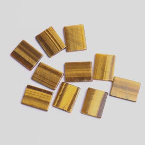 May include: A collection of rectangular tiger's eye gemstones. The stones have a polished finish, displaying bands of golden yellow and brown hues. The gemstones are arranged on a white surface, showcasing their natural patterns and colors.