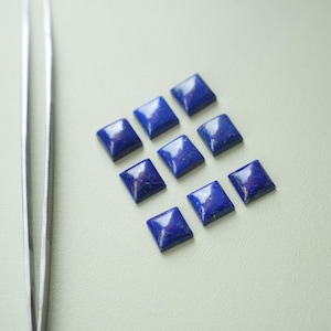 May include: Nine square-cut, deep blue lapis lazuli gemstones with a polished finish. The gemstones are arranged in a grid pattern on a light-colored surface. A pair of silver tweezers is visible on the left side of the image.