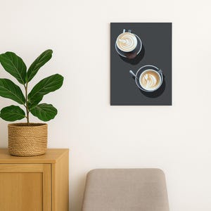 Puede incluir: Una impresión enmarcada con dos tazas de café con arte latte, vista desde arriba. Las tazas están sobre un fondo gris oscuro. La impresión cuelga de una pared blanca, con una planta en una cesta tejida y una lámpara.