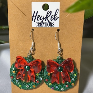May include: A pair of festive earrings featuring a green glitter wreath design with a red bow and a green gem in the center. The earrings are attached to silver hooks and are displayed on a brown card with the text "HeyReb CREATIONS".