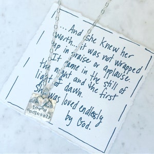 Puede incluir: Un collar de plata con un colgante cuadrado que dice "loved endlessly" y que presenta un par de alas de ángel y un corazón. El collar se muestra en un trozo de papel con texto escrito a mano que dice: "...And she knew her worth... it was not wrapped up in praise or applause. It came in the still of the night and the first light of dawn. She was loved endlessly by God."